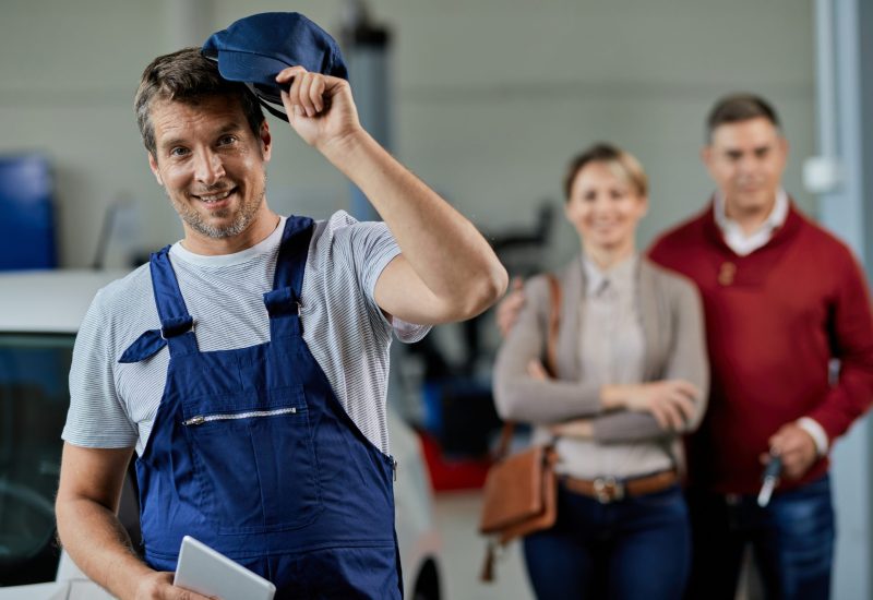 happy auto mechanic taking his hat while standing workshop looking camera his customers are background scaled happy auto mechanic taking his hat while standing workshop looking camera his customers are background scaled
